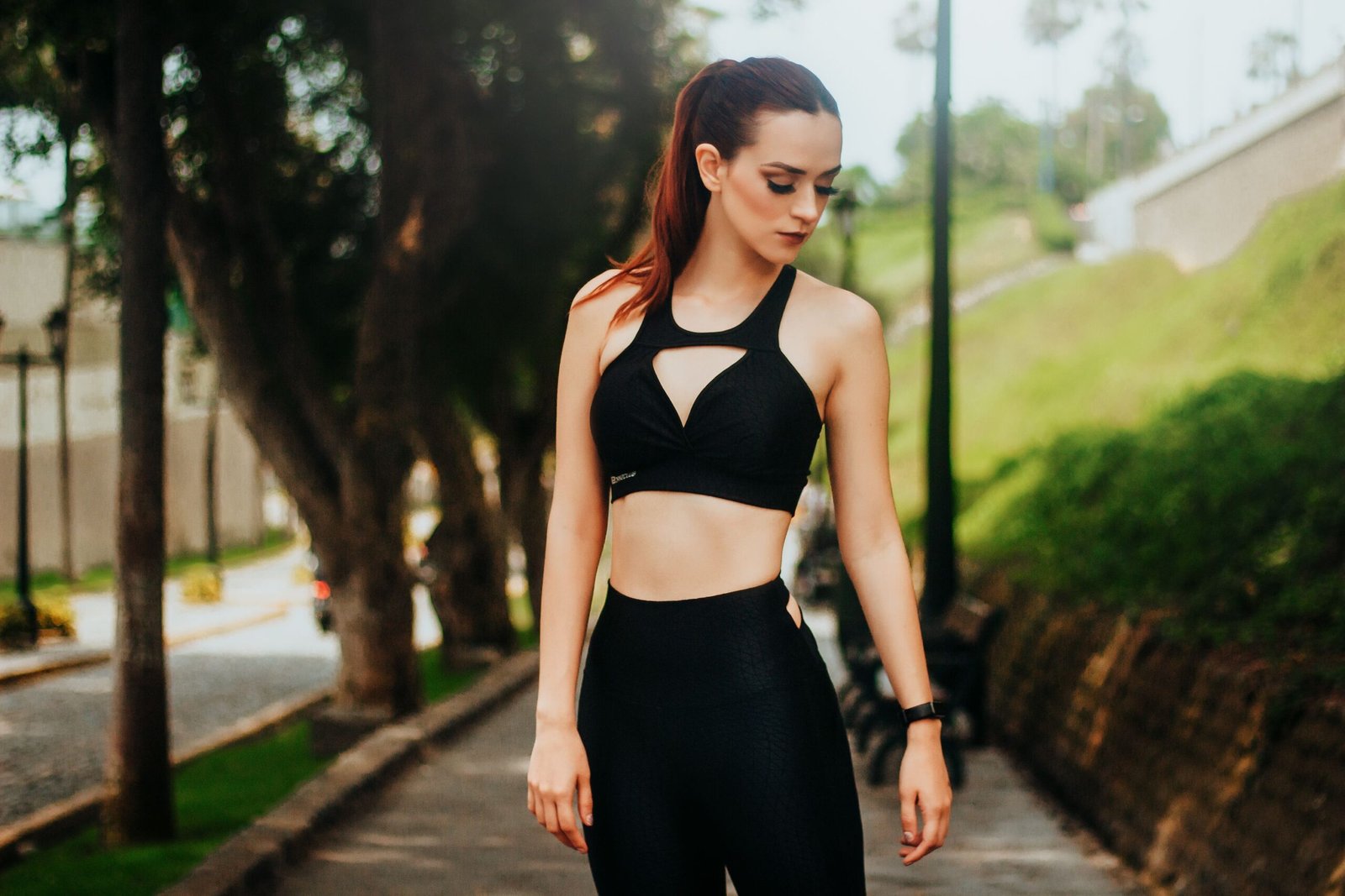 pexels photo 7862954 7862954 Calm young female wearing black leggings and top standing on walkway in green park and looking down in sunlight